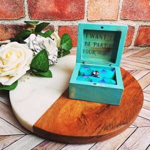 A wooden ring box or a wooden jewelry box stained in aqua, with silver vinyl saying I want to be part of your world. A cushion is wrapped in mermaid scale fabric where a ring or necklace can sit. There are built in barrel hinges and a magnetic latch.