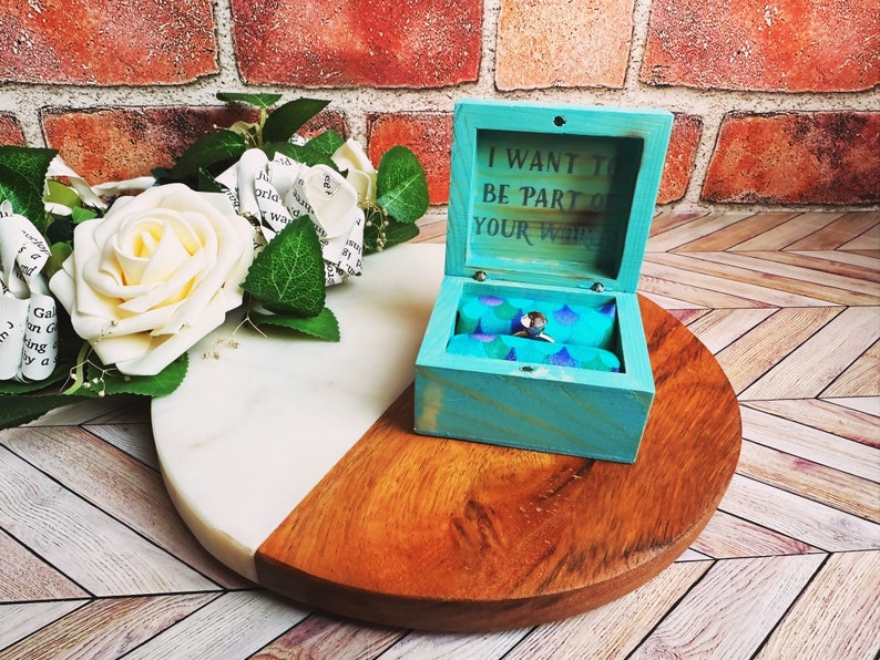 A wooden ring box or a wooden jewelry box stained in aqua, with silver vinyl saying I want to be part of your world. A cushion is wrapped in mermaid scale fabric where a ring or necklace can sit. There are built in barrel hinges and a magnetic latch.