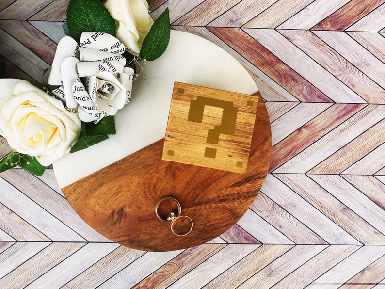 A wooden ring box or a wooden jewelry box stained in a honey color, with gold vinyl in the shape of an 8-bit question mark.