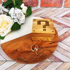 A wooden ring box or a wooden jewelry box stained in a honey color, with gold vinyl in the shape of an 8-bit question mark.