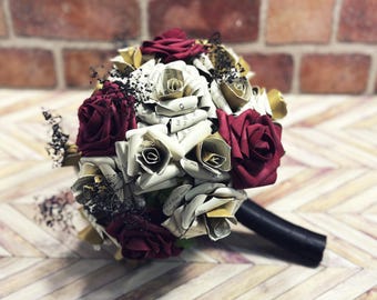 May include: A bouquet featuring deep burgundy fabric roses, gold-toned paper flowers, and roses crafted from printed book pages. The arrangement includes sprigs of dark, dried baby's breath and is finished with a solid black handle at the base.