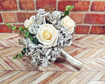 May include: A bouquet featuring cream-colored foam roses interspersed with handmade paper flowers crafted from printed book pages. The arrangement includes sprigs of green eucalyptus and dried brown filler, with a handle wrapped in smooth cream ribbon.