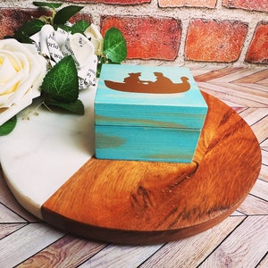 A wooden ring box or a wooden jewelry box stained in a beautiful aqua color, with gold vinyl spelling showing the couple during their first moment together.