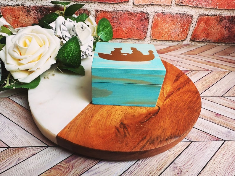 A wooden ring box or a wooden jewelry box stained in a beautiful aqua color, with gold vinyl spelling showing the couple during their first moment together.