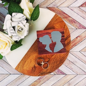 May include: A circular wooden display board with a white and natural wood finish, featuring a small rectangular box with a grey silhouette of a couple facing each other. Two gold-toned rings rest on the board, accompanied by cream-colored artificial roses.