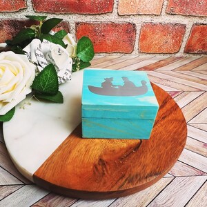 A wooden ring box or a wooden jewelry box stained in a beautiful aqua color, with silver vinyl spelling showing the couple during their first moment together.