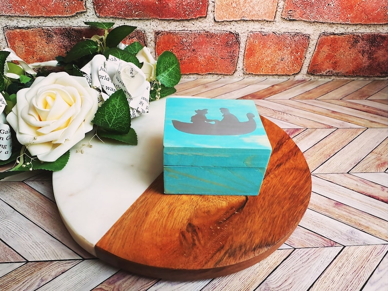 A wooden ring box or a wooden jewelry box stained in a beautiful aqua color, with silver vinyl spelling showing the couple during their first moment together.