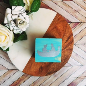 A wooden ring box or a wooden jewelry box stained in a beautiful aqua color, with silver vinyl spelling showing the couple during their first moment together.