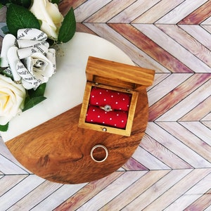 A wooden ring box or a wooden jewelry box stained in a honey color. Inside is a cushion wrapped in red fabric with white dots where a ring or necklace can nestle. You can see built in barrel hinges and a magnetic latch to keep your box closed.