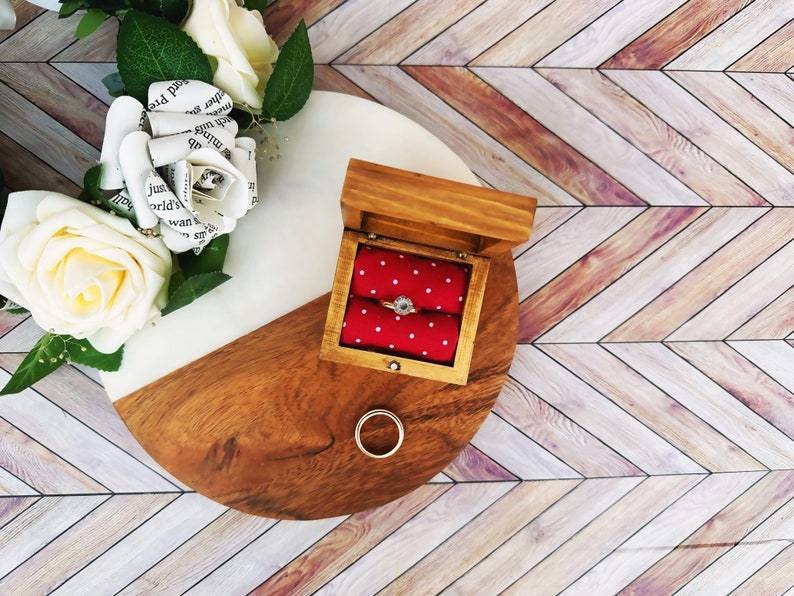 A wooden ring box or a wooden jewelry box stained in a honey color. Inside is a cushion wrapped in red fabric with white dots where a ring or necklace can nestle. You can see built in barrel hinges and a magnetic latch to keep your box closed.