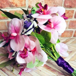 A hand tied bouquet made with pink orchids, book page roses layered with purple cardstock, pink foam roses, faux eucalyptus picks and fern greens. The bouquet has exposed stems and is tied with a matching purple ribbon.