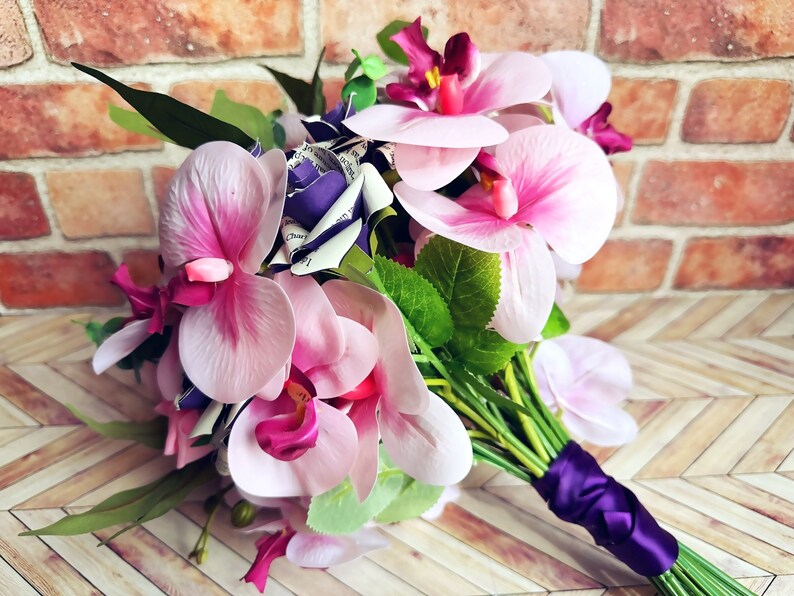 A hand tied bouquet made with pink orchids, book page roses layered with purple cardstock, pink foam roses, faux eucalyptus picks and fern greens. The bouquet has exposed stems and is tied with a matching purple ribbon.