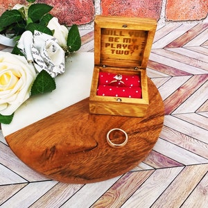 A wooden ring box or a wooden jewelry box stained in a honey color, with gold vinyl saying will you be my player two. A cushion is wrapped in red fabric with white dots where a ring or necklace can sit.