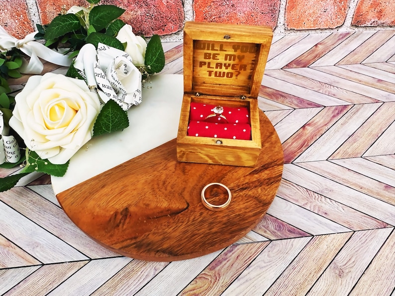 A wooden ring box or a wooden jewelry box stained in a honey color, with gold vinyl saying will you be my player two. A cushion is wrapped in red fabric with white dots where a ring or necklace can sit.