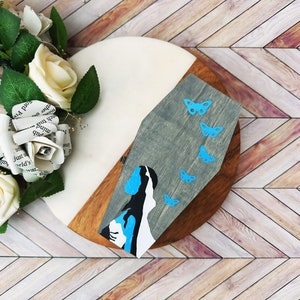 May include: A wooden coffin-shaped box with a blue and white painted image of a woman with butterflies on a gray background.