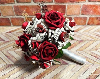 May include: A bouquet featuring deep red foam roses interspersed with handmade paper flowers crafted from printed book pages. The arrangement includes small sprigs of dried baby's breath, green fabric leaves, and a handle wrapped in white satin ribbon.