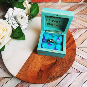 A wooden ring box or a wooden jewelry box stained in a vivid aqua color. Inside is a cushion wrapped in mermaid scale fabric where a ring or necklace can nestle. You can see built in barrel hinges and a magnetic latch to keep your box closed.