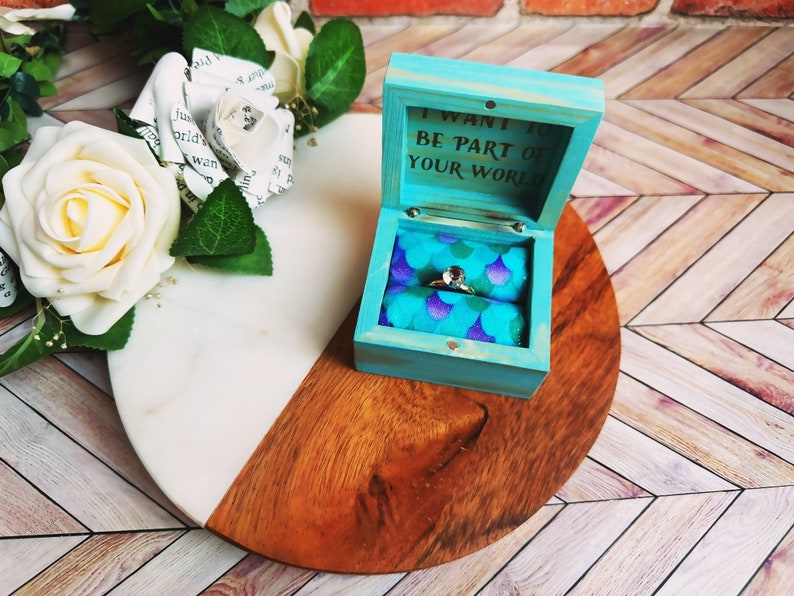 A wooden ring box or a wooden jewelry box stained in a vivid aqua color. Inside is a cushion wrapped in mermaid scale fabric where a ring or necklace can nestle. You can see built in barrel hinges and a magnetic latch to keep your box closed.