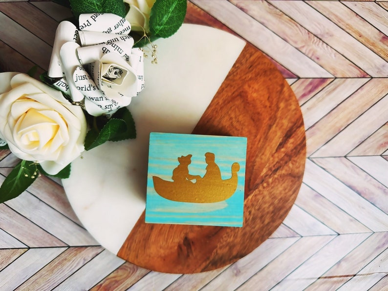 A wooden ring box or a wooden jewelry box stained in a vivid aqua color, with gold vinyl spelling showing the couple during their first moment together.