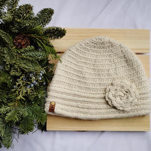 Op de afbeelding: Een crèmekleurige gebreide beanie met een gehaakte bloemversiering. De beanie heeft een lederen label met de tekst "City Chic" erop.