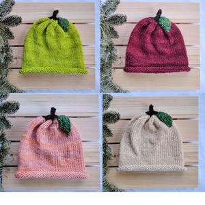 May include: Four knitted beanies in shades of green, burgundy, pink, and beige. Each hat features a brown stem and a green leaf accent, resembling a fruit. The hats are displayed on a wooden surface, showcasing the handmade quality.