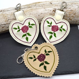 May include: Three heart-shaped leather keychains with embroidered roses and green leaves. The keychains are white, beige, and cream.