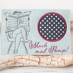 Pode incluir: Uma base de feltro azul claro com um design bordado. O design apresenta uma pessoa lendo um jornal com o texto "Horoskop Urlaubsblatt" e um círculo azul marinho com bolinhas brancas. O texto "Mach mal Pause!" também é bordado.