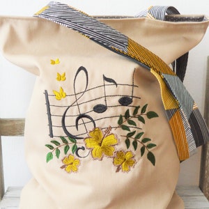Könnte beinhalten: Eine beige Canvas-Tragetasche mit einem schwarz-gelb gestreiften Riemen. Die Tasche hat ein gesticktes Design mit Noten, einem Violinschlüssel und gelben Blumen.