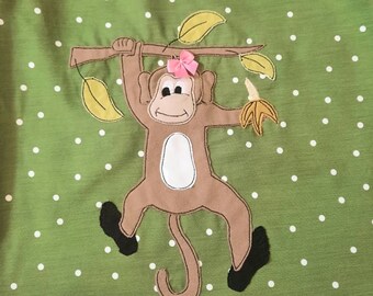 May include: Green fabric with white polka dots featuring an appliqued monkey design. The monkey is brown with a white belly, black feet, and a pink bow. It hangs from a branch with yellow leaves and holds a banana.