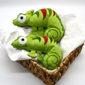 May include: Two plush green chameleon toys with red and black accents, resting in a woven basket lined with white fabric. The chameleons have large, round eyes and are designed with embroidered details.