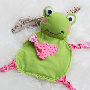 May include: Green frog-shaped plush toy with a pink heart and pink and white gingham bows. The toy is made of soft fabric and is suitable for babies and toddlers.