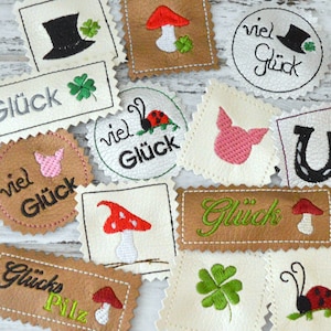 May include: A collection of embroidered iron-on patches featuring various good luck charms and the German word "Glück" (meaning "luck") in different styles. The charms include a four-leaf clover, a horseshoe, a ladybug, a mushroom, a pig, and a top hat.