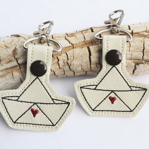 May include: Two white leather keychains with a paper boat design and a red heart in the center. The keychains are attached to silver key rings.