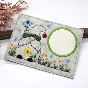 May include: Embroidered felt gnome with a red hat, green beard, and yellow flower. The gnome is surrounded by green grass, blue butterflies, and orange flowers. A white circle with a green outline is on the right side of the image.