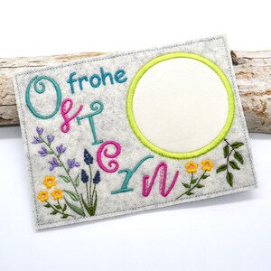 Puede incluir: Una tarjeta de Pascua de fieltro gris con texto bordado y detalles florales. La tarjeta presenta la frase alemana "frohe Ostern" en turquesa y rosa, un marco circular y flores bordadas.