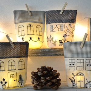 Könnte beinhalten: Fünf weiße Stoff-Haus-förmige Lichtbezüge mit gestickten Details, darunter Fenster, Türen und ein Baum. Die Bezüge hängen an einem Holzregal mit einer Kiefernzapfen in der Mitte.