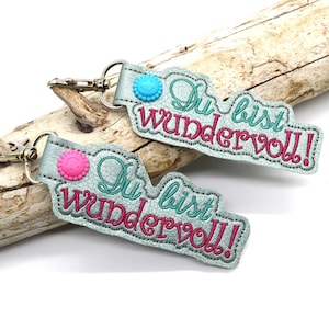 May include: Two light blue keychains with a smiling face button and the text "Du bist wundervoll!" in pink and teal embroidery.
