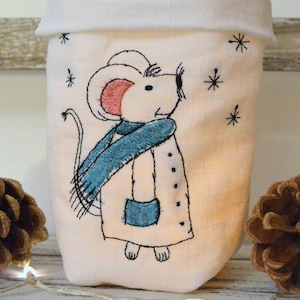 Op de afbeelding: Een witte stoffen tas met een handgestikt ontwerp van een muis die een blauwe sjaal en jas draagt. De muis kijkt omhoog naar sneeuwvlokken.
