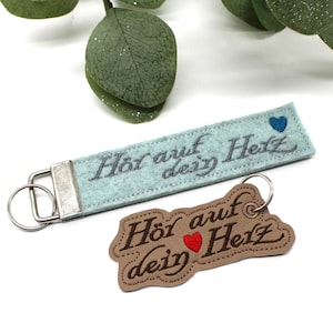 May include: Two keychains with the German text "Hör auf dein Herz" (Listen to your heart). One is a light blue felt strap with a silver metal clasp and a blue heart. The other is a brown leather-like tag with a red heart.