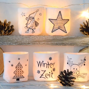 May include: Five white fabric candle holders with black embroidered designs. The designs include a star, a Christmas tree, a reindeer, a snowman, and the text "Winter Zeit".