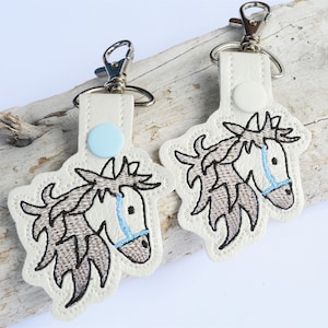 May include: Two white leather keychains with embroidered horse heads. The horses have brown manes and tails and blue bridles. The keychains have silver key rings.