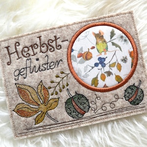 Könnte beinhalten: Eine beige Filzkarte mit dem Text "Herbst-geflüster", der in braunem Garn gestickt ist. Die Karte zeigt ein kreisförmiges gesticktes Patch mit weißem Hintergrund und einer braunen Maus mit gelber Weste, die auf einem Zweig mit Beeren und Blättern steht. Die Karte zeigt auch zwei gestickte braune und grüne Kastanien.