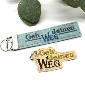 May include: Two keychains with the German phrase "Geh deinen Weg" embroidered on them. One is a light blue rectangular keychain with silver hardware. The other is a beige keychain in the shape of a key.