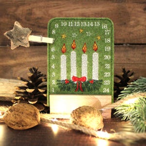 May include: A green felt Advent calendar with embroidered white candles, red flames, and a red bow. Numbers 1-24 are on the side. A wooden star is held by a clothespin. Pinecones and walnuts are in the foreground.