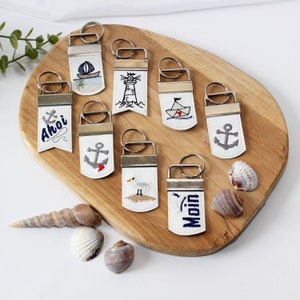 May include: Nine white leather keychains with different nautical themed embroidery designs. The designs include an anchor, a lighthouse, a sailboat, a seagull, and the words "Ahoi" and "Moin".
