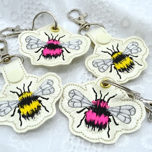 May include: Four keychains with embroidered bumblebees. Two bumblebees are pink and black, and two are yellow and black. The keychains are made of white leather and have silver key rings.