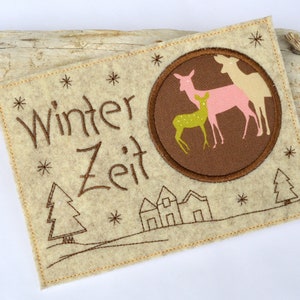 May include: A beige felt ornament with brown embroidery depicting a winter scene with a family of deer, a small village, and the text "Winter Zeit".