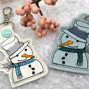 May include: Two snowman keychains, one white with a blue scarf and one light blue with a teal scarf. Both snowmen have black eyes, a carrot nose, and a gray hat.