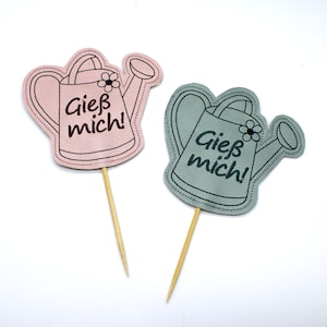May include: Two watering can-shaped decorative toppers. One is pink, the other is gray-green. Each has the German text "Gieß mich!" and a small flower. They are attached to wooden sticks, suitable for cakes or plants.