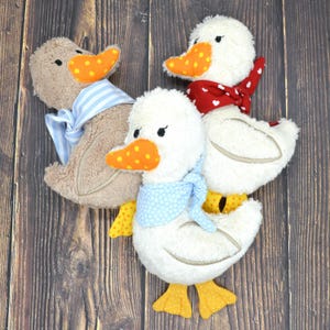 May include: Three plush duck toys with orange beaks and yellow feet. One duck is brown with a blue and white striped scarf. The other two are white, one with a red heart-patterned scarf and the other with a blue polka-dot scarf.